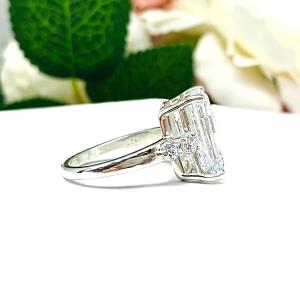 A sterling silver engagement ring featuring a stunning 14x10mm emerald cut cubic zirconia center stone flanked by two sparkling round CZ accents on each side. Shown on a white surface with a pastel floral background.