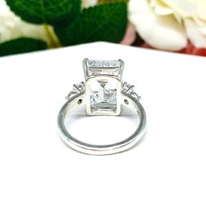 A sterling silver engagement ring featuring a stunning 14x10mm emerald cut cubic zirconia center stone flanked by two sparkling round CZ accents on each side. Shown on a white surface with a pastel floral background.
