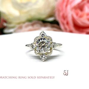 A silver ring with a large round cz surrounded by smaller czs. The ring has a delicate design with a scalloped edge. The ring is on a white surface with a blurry image of pink flowers in the background. The text 'MATCHING RING SOLD SEPARATELY' is at the bottom of the image. The letters 'SJ' are in the bottom right corner of the image.