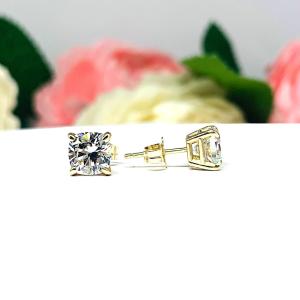 A pair of yellow gold stud earrings with brilliant cushion cut clear cubic zirconia stones. The basket style soft claw prong earrings are on a white surface with the left earring facing forward to showcase the brilliant faceting of the czs and the right earring facing to the right to display the center post and butterfly friction clasp.