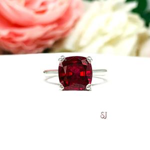 Sterling silver ring with a cushion cut lab created deep red ruby set in a split-prong setting set against a white surface with soft-focus pink and white flowers in the background.