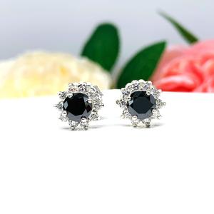 A pair of sterling silver stud earrings set with round black moissanites with several small colorless moissanites surrounding them in a halo. The earrings are shown on a white surface with a background of soft-focus pink and white flowers.