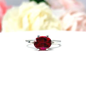 Sterling silver ring with a horizontally set (east/west) oval lab created deep red ruby set in a 6-prong setting set against a white surface with soft-focus pink and white flowers in the background.