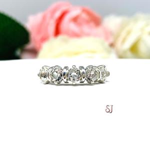 A sterling silver ring featuring five 4mm round white topaz gemstones in a row. The ring has rounded bars separating each stone. Displayed on a white surface with pastel floral background.