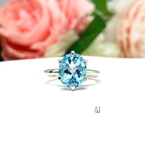 A sterling silver 6-prong ring featuring a 10x8mm oval saturated tone sky blue topaz gemstone. Shown on a white surface with a pastel floral background.