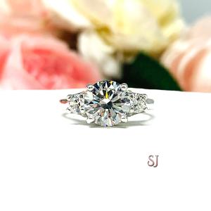 Sterling silver 3-stone engagement ring featuring an 8mm round brilliant cut near colorless cubic zirconia center stone with two smaller round CZ side stones.  Displayed on a white surface with a soft floral background.