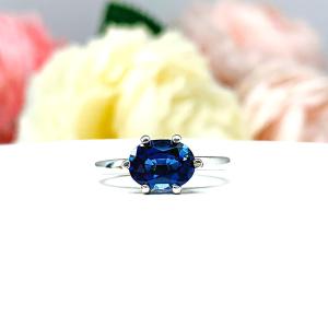 Sterling silver ring with a horizontally set (east/west) oval lab created blue sapphire set in a 6-prong setting set against a white surface with soft-focus pink and white flowers in the background.
