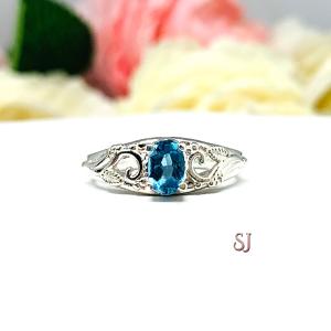 A sterling silver ring with a 6x4mm oval medium blue tone London blue topaz gemstone set in a delicate filigree design. Displayed on a white surface with a pastel floral background. The letters 'SJ' appear in the bottom right corner.