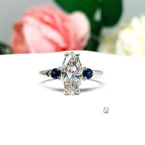 Stunning marquise-cut near colorless CZ engagement ring in sterling silver, accented with two round lab-created blue sapphires. Shown in size 7, the ring is nestled against a soft floral backdrop, radiating elegance and brilliance.