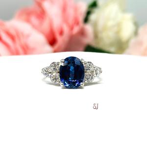 A sterling silver ring with an oval lab blue sapphire surrounded by smaller round czs. The ring is set on a white surface with pink and white flowers in the background. The letters 'SJ' appear in the bottom right corner.