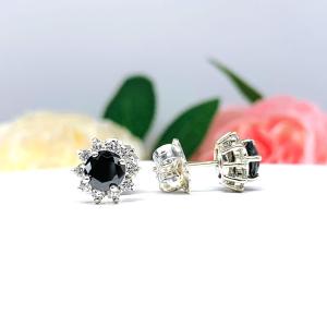 A pair of sterling silver stud earrings set with round black moissanites with several small colorless moissanites surrounding them in a halo. The earrings are shown on a white surface with a background of soft-focus pink and white flowers.