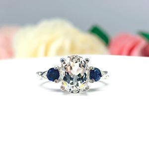 A sterling silver ring with an oval natural white topaz center and round iolite accents. The ring, with its twist milgrain design and contrasting colors is striking set against a background of pink and white flowers.