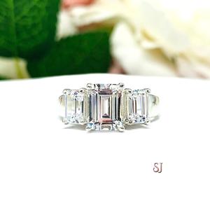 Sterling silver 3-stone engagement ring set with an 8x6mm emerald cut cubic zirconia and flanked by a 6x4mm emerald cut cz on each side. The ring is resting on a white surface with a background of pink and white flowers.