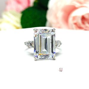 A sterling silver engagement ring featuring a stunning 14x10mm emerald cut cubic zirconia center stone flanked by two sparkling round CZ accents on each side. Shown on a white surface with a pastel floral background.
