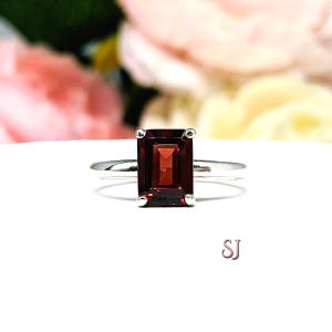 A sterling silver solitaire ring set with an emerald cut earthy red Mozambique garnet gemstone. Shown displayed on a white surface with a pastel floral background.