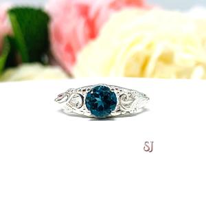 A sterling silver ring with a richly saturated 6mm round London blue topaz gemstone set in a delicate filigree design. Displayed on a white surface with a pastel floral background. The letters 'SJ' appear in the bottom right corner.
