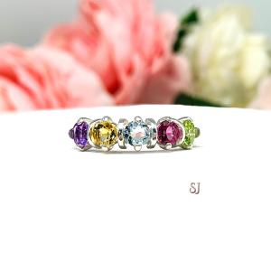 A sterling silver "spring season" ring with five gemstones. The 4mm round gemstones are purple (amethyst), yellow (citrine), blue (sky blue topaz), pink (pink topaz), and green (peridot). Displayed on a white surface with pastel floral background.