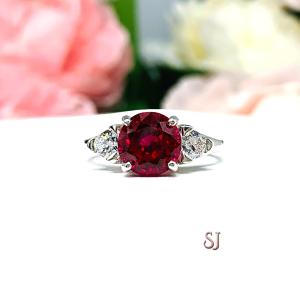 A sterling silver three stone ring featuring a deep red 8mm round cut lab created ruby flanked by a 4mm trillion cz on each side. The ring is resting on a white surface with soft-focus pink and white flowers in the background.