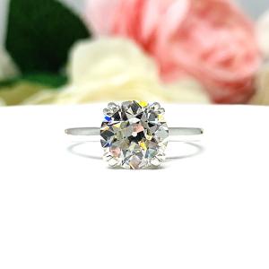A romantic close-up of a 9mm (3 ct) old European cut near-colorless CZ solitaire engagement ring. Set in a sterling silver split prong cathedral-style mount, the ring gleams with vintage-inspired elegance. Soft pink and white flowers blur in the background, adding a dreamy, bridal ambiance.