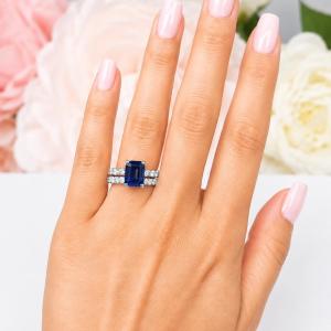 A woman's hand wearing a sterling silver engagement ring featuring a 10x8mm emerald cut blue sapphire and 2.5mm round czs on the band. Shown with matching wedding band. Model image for size and styling reference — not a photo of the actual ring.
