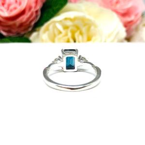 Close-up of an 8x6mm emerald-cut London Blue Topaz ring with moissanite side stones, set in a sterling silver twist band with milgrain detailing; soft floral background enhances the elegance.