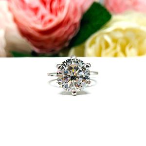 Close-up view of a 10mm near colorless (G/H color) round brilliant cut cubic zirconia solitaire engagement ring in a classic 6-prong setting. The setting is simple yet elegant, allowing the center stone to take center stage. The ring is displayed against a soft, blurred background of pastel-colored flowers, creating a romantic and delicate ambiance.