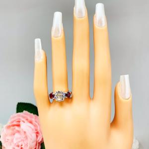 Mannequin hand with long white shimmery nails, displayed against a soft white-gray background with pink and white floral accents. The hand is wearing a sterling silver 3-stone ring with a 9mm near colorless round brilliant cut cubic zirconia center that radiates icy sparkle, flanked by two 5mm round deep red lab-created rubies.
