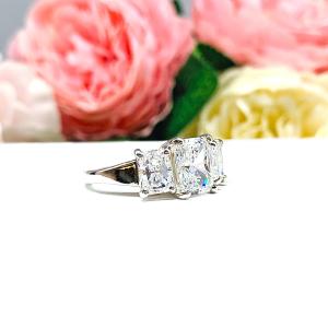 Sterling silver 3-stone engagement ring featuring a 9x7mm radiant cut cubic zirconia center stone with two 7x5mm radiant CZ side stones. The highly polished wide band tapers for comfort. Displayed on a white surface with soft floral background.