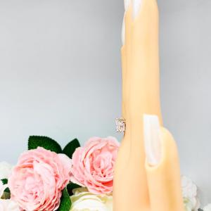 Mannequin hand with long white shimmery nails, displayed against a soft white-gray background with pink and white floral accents. The hand is wearing a sterling silver 3-stone engagement ring featuring a 9x7mm radiant cut cubic zirconia center stone with two 7x5mm radiant CZ side stones. The highly polished wide band tapers for comfort. Displayed on a white surface with soft floral background.
