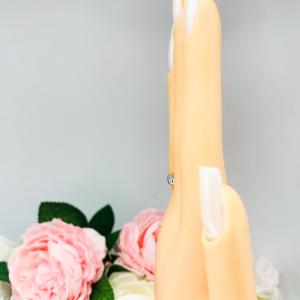 Mannequin hand with long white shimmery nails, displayed against a soft white-gray background with pink and white floral accents. The hand is wearing a sterling silver band featuring a 5×3 mm emerald‑cut green tourmaline set flush in a polished band.