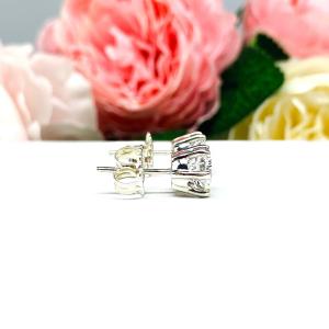 A pair of sterling silver six prong stud earrings set with round brilliant lab white sapphires. The earrings have center posts and butterfly clasps and are displayed on a white surface with a floral background.