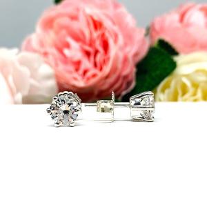 A pair of sterling silver six prong stud earrings set with round brilliant lab white sapphires. The earrings have center posts and butterfly clasps and are displayed on a white surface with a floral background.