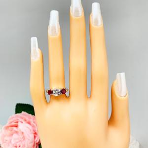 Mannequin hand with long white shimmery nails, displayed against a soft white-gray background with pink and white floral accents. The hand is wearing a sterling silver 3-stone ring with a 6mm round brilliant cut cubic zirconia center that radiates icy sparkle, flanked by two 5mm round deep red lab-created rubies.