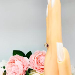 Mannequin hand with long white shimmery nails, displayed against a soft white-gray background with pink and white floral accents. The hand is wearing a sterling silver 3-stone ring with a 9mm near colorless round brilliant cut cubic zirconia center that radiates icy sparkle, flanked by two 5mm round deep red lab-created rubies.