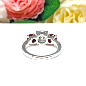 Sterling silver 3-stone ring with a round brilliant cut colorless (D color) cubic zirconia center flanked by two deep red lab-created rubies. The ring is set on a white surface with a background of pink and white flowers.
