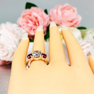Mannequin hand with long white shimmery nails, displayed against a soft white-gray background with pink and white floral accents. The hand is wearing a sterling silver 3-stone ring with a 9mm near colorless round brilliant cut cubic zirconia center that radiates icy sparkle, flanked by two 5mm round deep red lab-created rubies.