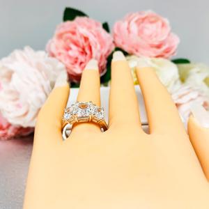 Mannequin hand with long white shimmery nails, displayed against a soft white-gray background with pink and white floral accents. The hand is wearing a sterling silver 3-stone engagement ring featuring a 9x7mm radiant cut cubic zirconia center stone with two 7x5mm radiant CZ side stones. The highly polished wide band tapers for comfort. Displayed on a white surface with soft floral background.