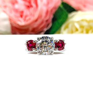 Sterling silver 3-stone ring with a round brilliant cut colorless (D color) cubic zirconia center flanked by two deep red lab-created rubies. The ring is set on a white surface with a background of pink and white flowers.