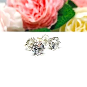A pair of sterling silver six prong stud earrings set with round brilliant lab white sapphires. The earrings have center posts and butterfly clasps and are displayed on a white surface with a floral background.