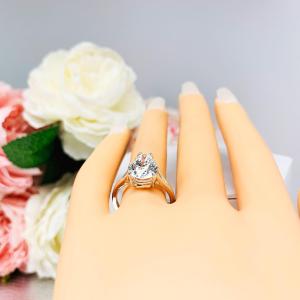 Mannequin hand with long white shimmery nails, displayed against a soft white-gray background with pink and white floral accents. The hand is wearing an elegant 4-prong sterling silver cathedral engagement ring set featuring a 13x9mm near colorless precision-cut pear cubic zirconia. Displayed on a white surface with a floral background.