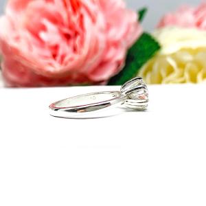 Sterling silver round-cut DEF moissanite 6-prong engagement ring. Displayed on a white surface with pink and white flowers in the background.