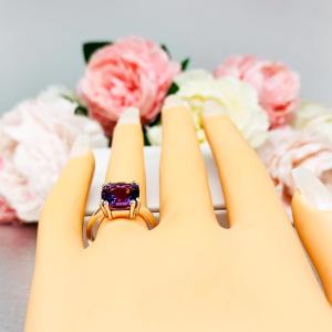 Mannequin hand with long white shimmery nails, displayed against a soft white-gray background with pink and white floral accents. The hand is wearing a sterling silver solitaire ring featuring a 10mm checkerboard cushion-cut vibrant African amethyst gemstone. The vibrant purple gem ring is set against a soft floral background that enhances the ring’s natural elegance.
