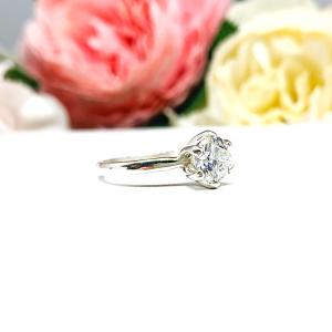 Sterling silver round-cut DEF moissanite 6-prong engagement ring. Displayed on a white surface with pink and white flowers in the background.
