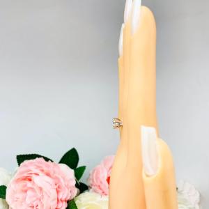 Mannequin hand with long white shimmery nails, displayed against a soft white-gray background with pink and white floral accents. The hand is wearing a sterling silver round-cut DEF moissanite 6-prong engagement ring. Displayed on a white surface with pink and white flowers in the background.