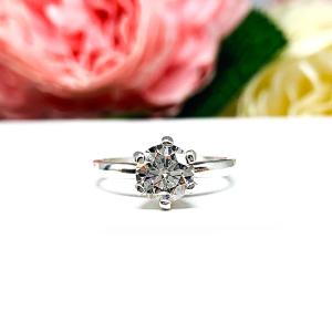 Sterling silver round-cut DEF moissanite 6-prong engagement ring. Displayed on a white surface with pink and white flowers in the background.