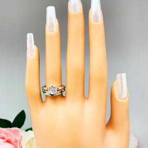 Mannequin hand with long white shimmery nails, displayed against a soft white-gray background with pink and white floral accents. The hand is wearing a sterling silver  five-stone curved wedding band with three 3mm round lab-created emeralds and two 3mm round DEF moissanites. Shown with a round moissanite engagement ring (not included).