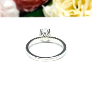 A solitaire engagement ring with a round brilliant DEF moissanite. The setting is a classic four-prong design, and the band is a simple, sterling silver band. The ring is displayed on a white background with blurred pink and yellow flowers in the background.