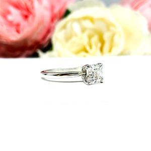 Sterling silver 4-prong asscher cut DEF moissanite solitaire engagement ring. The ring is resting on a white surface with a background of pink and white flowers.