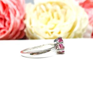 Sterling silver ring with an 8x6mm oval lab created padparadscha sapphire set in a 6-prong setting set against a white surface with soft-focus pink and white flowers in the background.