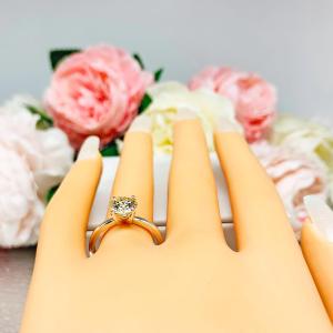 Mannequin hand with long white shimmery nails, displayed against a soft white-gray background with pink and white floral accents. The hand is wearing a solitaire engagement ring with a round brilliant light champagne moissanite. The setting is a classic four-prong design, and the band is a simple, sterling silver band.